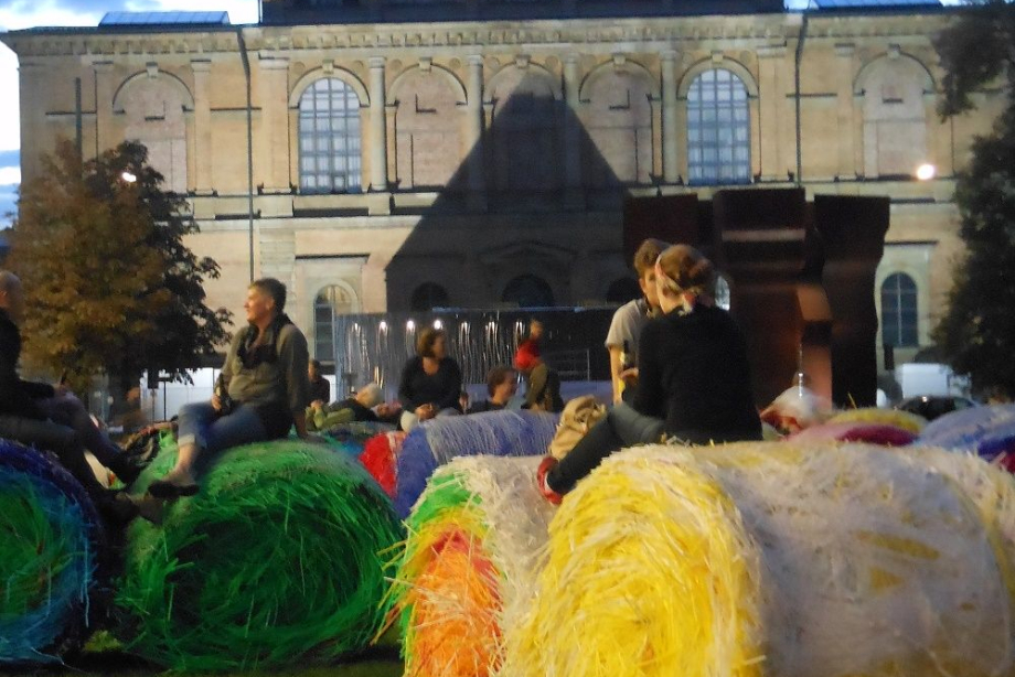In the evening sun, young people sit relaxed on top of the colored bales and chat. In the background the Alte Pinakothek, illuminated by the summer evening sun.