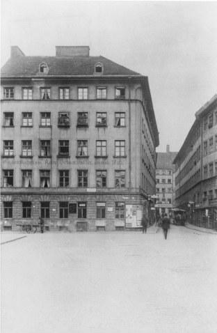 Auf einer historischen Schwarz-Weiß-Fotografie ist ein vierstöckiges Wohnhaus zu sehen. Im Erdgeschoss befindet sich das ehemalige Schwulenlokal „Schwarzfischer“. Man sieht Passanten und ein Pferdefuhrwerk und einen Mann mit Leiterwagen auf der Straße.