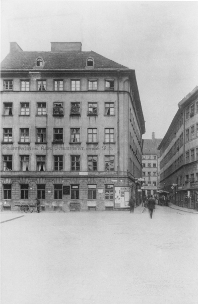 A historic black-and-white photograph shows a four-story apartment building. The former gay bar "Schwarzfischer" is located on the ground floor. One sees passers-by and a horse-drawn carriage and a man with a cart on the street.