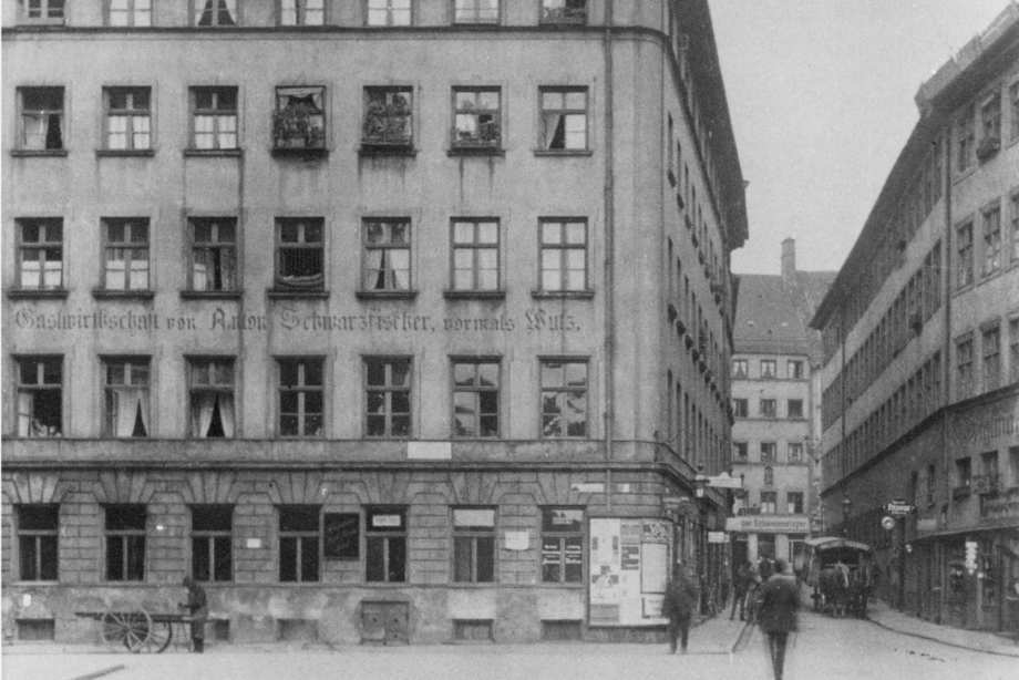 Auf einer historischen Schwarz-Weiß-Fotografie ist ein vierstöckiges Wohnhaus zu sehen. Im Erdgeschoss befindet sich das ehemalige Schwulenlokal „Schwarzfischer“. Man sieht Passanten und ein Pferdefuhrwerk und einen Mann mit Leiterwagen auf der Straße.