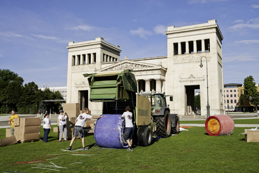 A tractor with a harvest trailer on a meadow on Königsplatz presses red and blue straw into large bales. Two young people help. In the background you can see the Propylaea.
