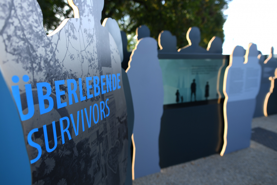 Blick auf eine Gruppe von Figuren-Silhouetten, links im Vordergrund die Aufschrift 'Überlebende', rechts unscharf im Hintergrund der Infoscreen zwischen den Figuren