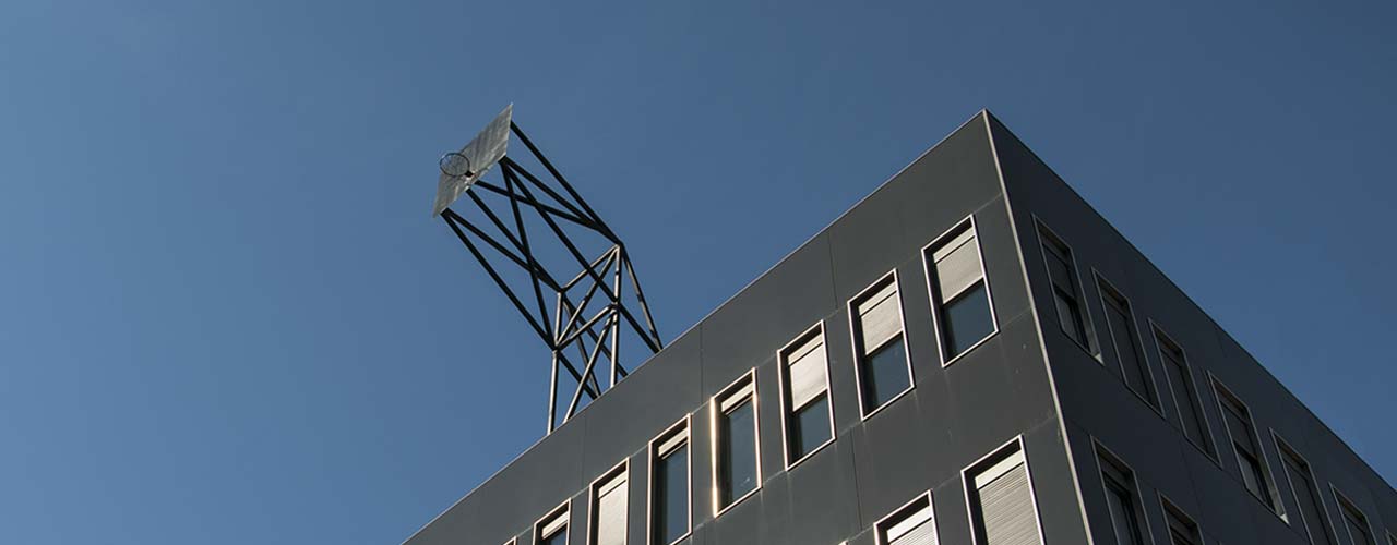 A basketball backboard is mounted on the roof of a tall building, jutting into the blue summer sky.