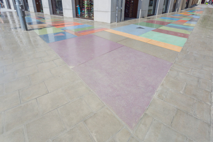 You can see a floor mosaic made of colored concrete blocks embedded in a sidewalk. There is a black chevron on one of the colored stones, and a pink chevron on another.
