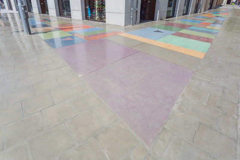 You can see a floor mosaic made of colored concrete blocks embedded in a sidewalk. There is a black chevron on one of the colored stones, and a pink chevron on another.