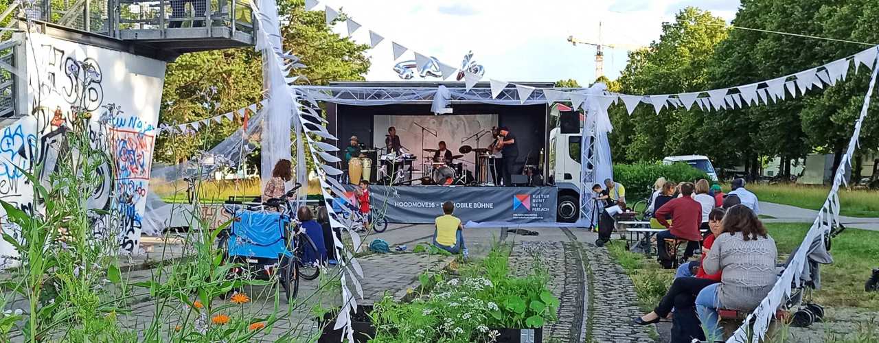 Die Musikkapelle „Lucile and the Rakibuam“ auf einer mobilen Bühne, die mit Hochzeitsschmuck verziert ist. Im Vordergrund sitzen Zuhörer*innen im Grünen.