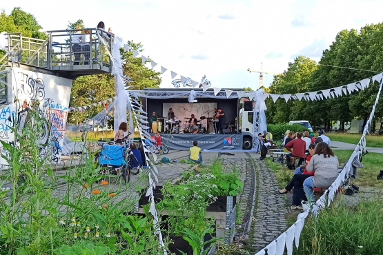 Die Musikkapelle „Lucile and the Rakibuam“ auf einer mobilen Bühne, die mit Hochzeitsschmuck verziert ist. Im Vordergrund sitzen Zuhörer*innen im Grünen.