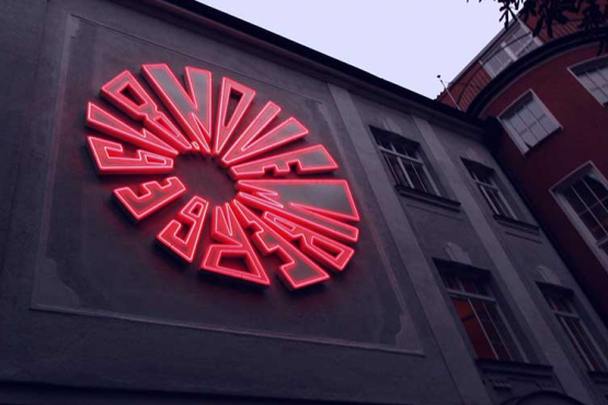 At dusk, an installation of bright red neon tubes appears on a house wall in the shape of a bomb detonation. The diameter of the installation is about 5 meters.