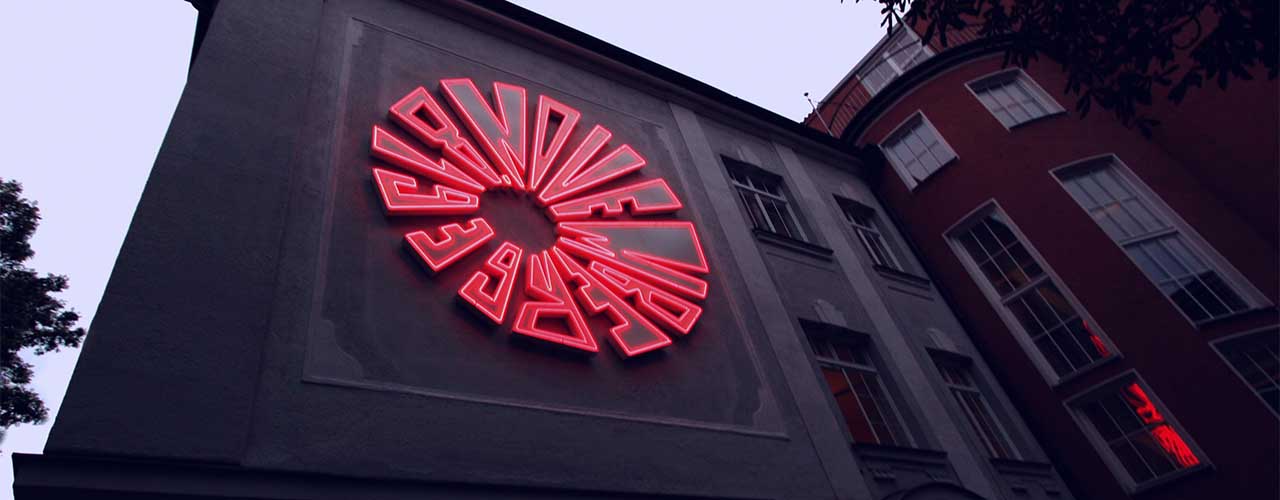 At dusk, an installation of bright red neon tubes appears on a house wall in the shape of a bomb detonation. The diameter of the installation is about 5 meters.