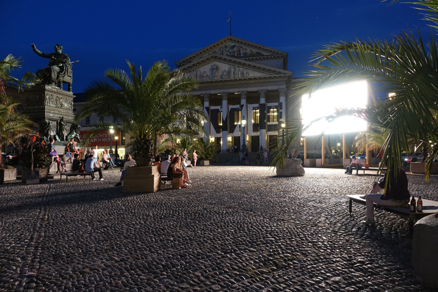 Night view of Max-Josef-Platz. In the background is a luminous display, in front people are sitting on seats between palm trees.