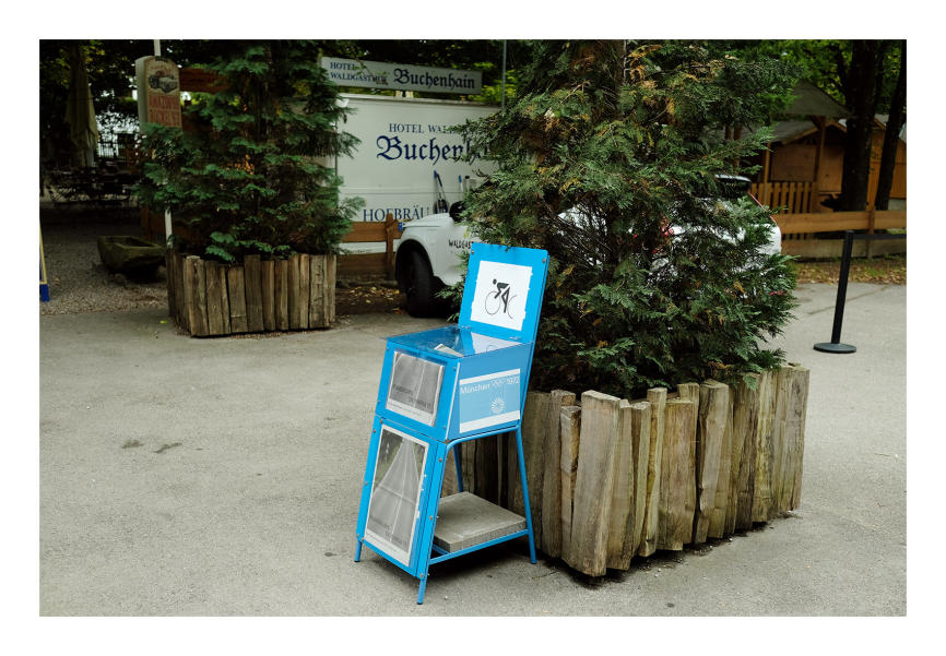 Blue newspaper box in front of a large plant trough, seen from diagonally right, another plant trough in the background, parked cars behind it.