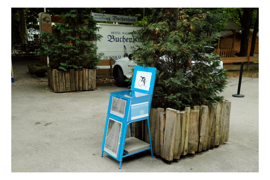 Blue newspaper box in front of a large plant trough, seen from diagonally right, another plant trough in the background, parked cars behind it.