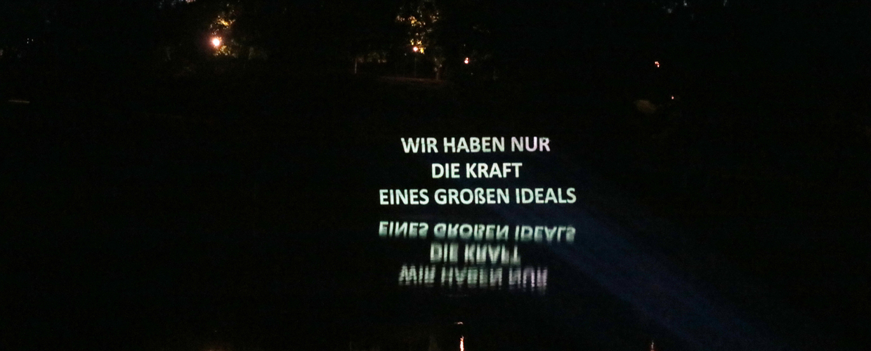 the sentence 'Wir haben nur die Kraft eines großen Ideals' is projected in a nocturnal setting against the background of trees, the projection is reflected in the water