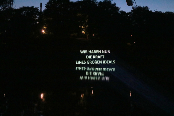 the sentence 'Wir haben nur die Kraft eines großen Ideals' is projected in a nocturnal setting against the background of trees, the projection is reflected in the water