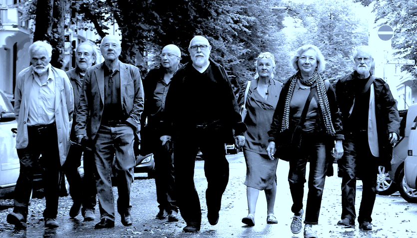 auf dem Schwarz-Weiß-Foto sind mehrere ältere Personen zu sehen, die auf regennasser Straße auf den/die Betrachter*in zulaufen