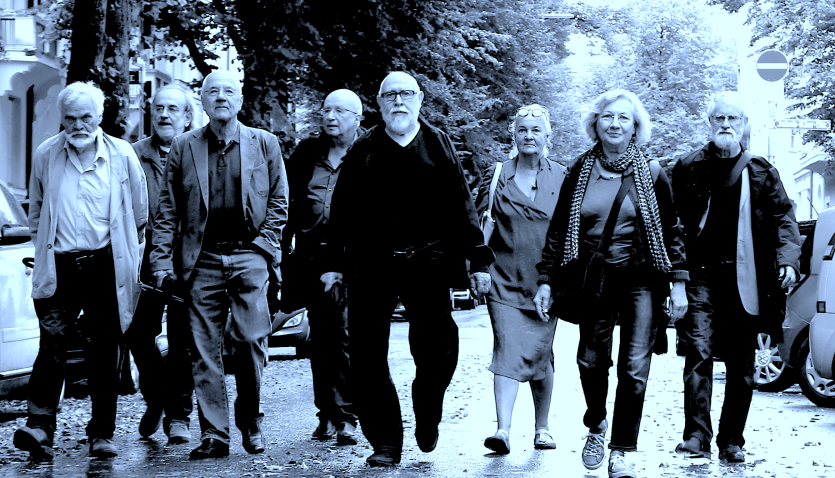 The black-and-white photo shows several elderly people walking towards the viewer on a rain-soaked street