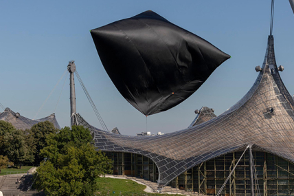A balloon-like sculpture made of thin black fabric can be seen flying over the Olympic Stadium. The sun shines brightly from the deep blue sky.