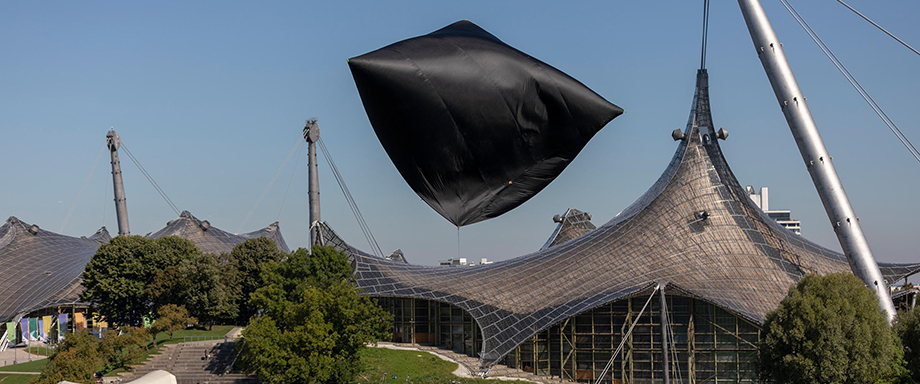 A balloon-like sculpture made of thin black fabric can be seen flying over the Olympic Stadium. The sun shines brightly from the deep blue sky.