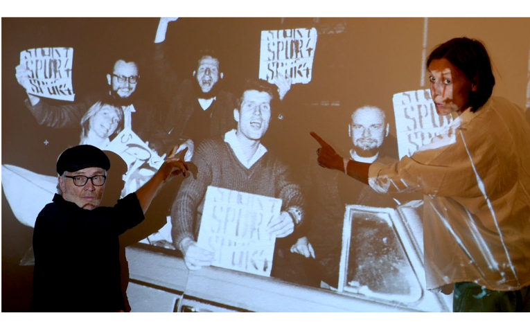 Rudolf Herz und Julia Wahren zeigen auf eine Projektion eines historischen Fotos, welches mehrere Personen mit Schildern zeigt
