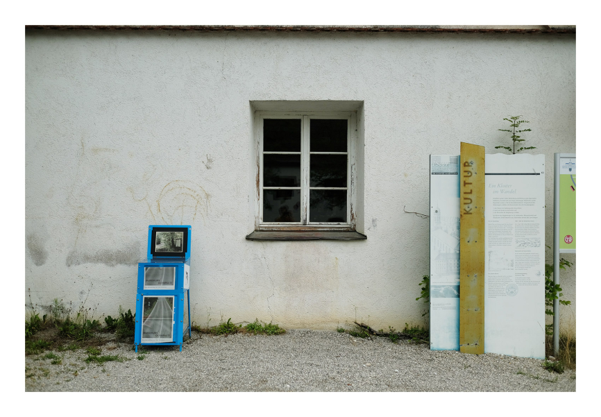 in front of a house wall with a mullioned window in the middle, to the left of it is a blue newspaper box, to the right of the window is an information board