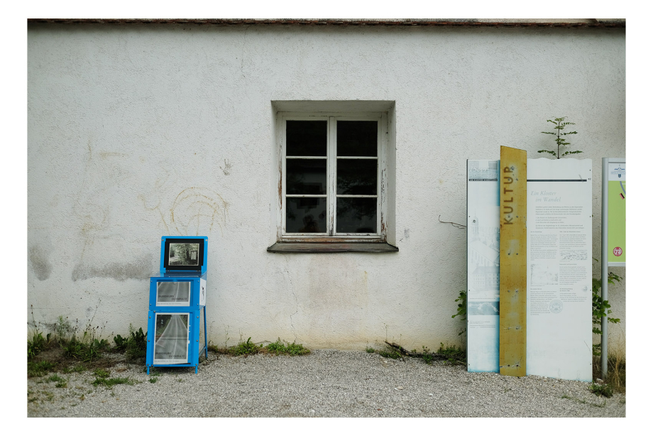 in front of a house wall with a mullioned window in the middle, to the left of it is a blue newspaper box, to the right of the window is an information board