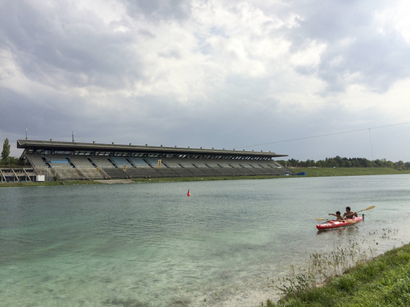 View of the Olympic regatta course in front and the empty grandstand against a cloudy sky, two people in a red two-man kayak in the foreground on the right.