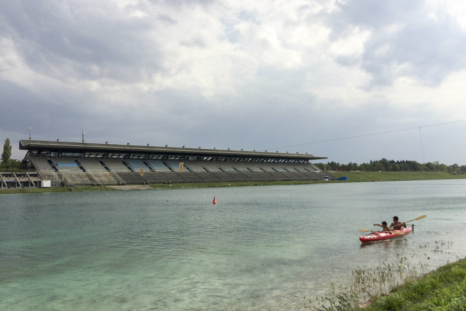 View of the Olympic regatta course in front and the empty grandstand against a cloudy sky, two people in a red two-man kayak in the foreground on the right.