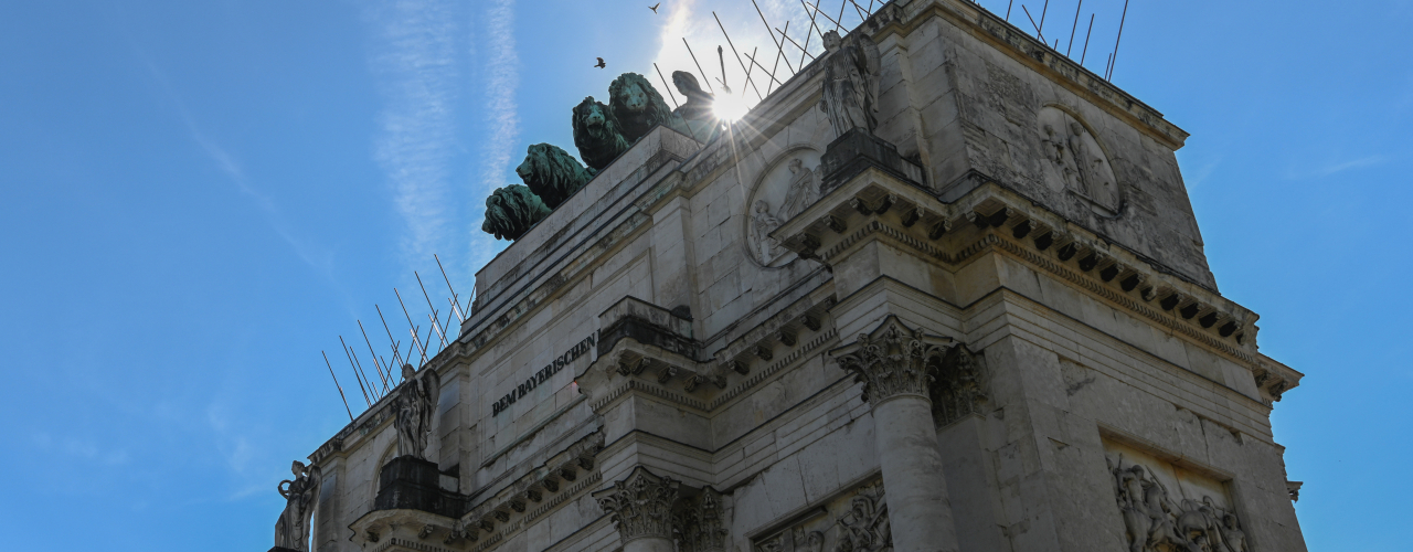 Blick von seitlich unten am Siegestor hoch, die Sonne blitzt hinter der Bavaria oben auf dem Siegestor hervor. Gegen den blauen Himmel sind die überdimensionierten Tauben Spikes zu sehen