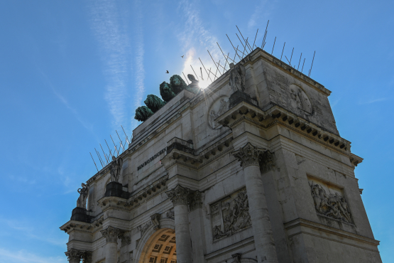 Blick von seitlich unten am Siegestor hoch, die Sonne blitzt hinter der Bavaria oben auf dem Siegestor hervor. Gegen den blauen Himmel sind die überdimensionierten Tauben Spikes zu sehen