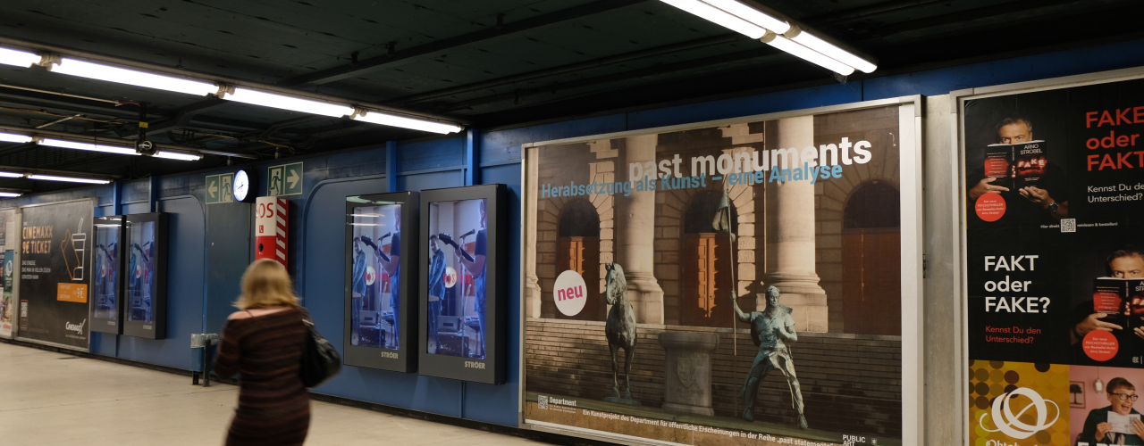 An underpass, various posters on the walls, the poster in the middle shows an equestrian statue, but horse and rider are standing to the left and right of the base