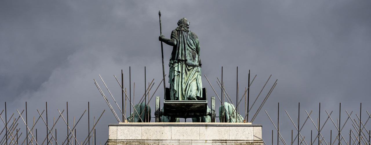 Blick auf den obersten Teil des Siegestors mit der Bavaria auf ihrem Wagen, umringt von den überdimensionalen Tauben Spikes, das Ganze vor einem gewittrig dunklen Himmel