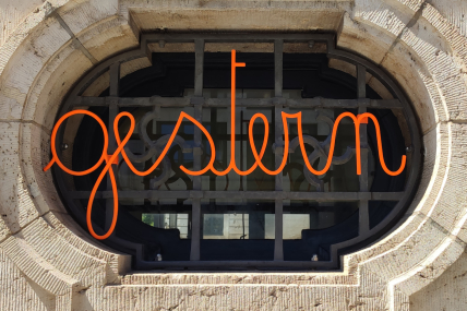 Orangefarbener Schriftzug "gestern" vor einem Fenster, auf dem sich das Fresko eines Hakenkreuzes befindet.