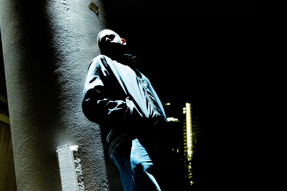nächtliches Foto eines Jugendlichen, der an eine Wand gelehnt steht, das Gesicht teils im Licht, teils beleuchtet, ist nicht erkennbar