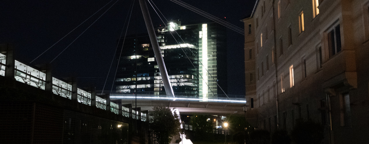 nächtliche Stadtszene bestehend aus einem Bürokomplex und einem Wohngebäude, jeweils mit teilweise beleuchteten Fenstern, sowie einer Brücke