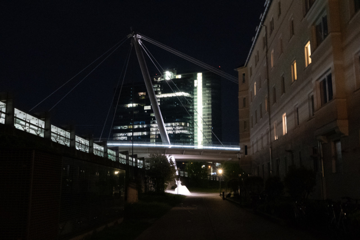 nächtliche Stadtszene bestehend aus einem Bürokomplex und einem Wohngebäude, jeweils mit teilweise beleuchteten Fenstern, sowie einer Brücke