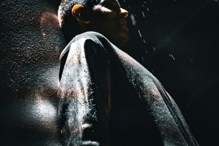 Night-time shot from the side of a young person leaning against a wall, detail of the upper part of the body, the face partly in the light, partly in the shadow.