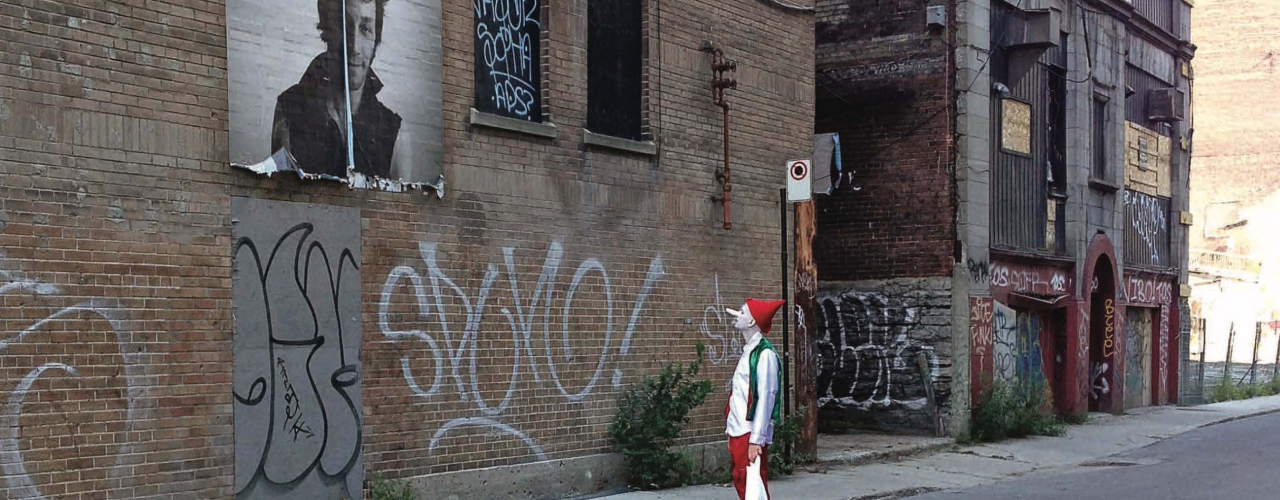 Das Bild zeigt den Künstler als Clown mit weiß geschminktem Gesicht, rotem Hut und in Kostüm in einer verlassenen Straße, vertieft in die Betrachtung eines übergroßen Porträts einer unbekannten Person.
