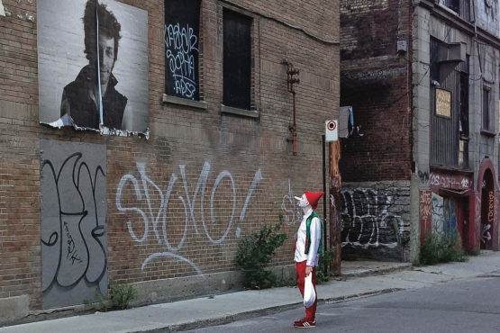 Image shows the artist dressed as a clown with a white-painted face, a red hat, and in costume in a deserted street, engrossed in the contemplation of an oversized portrait of an unfamiliar person.