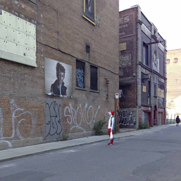 Image shows the artist dressed as a clown with a white-painted face, a red hat, and in costume in a deserted street, engrossed in the contemplation of an oversized portrait of an unfamiliar person.