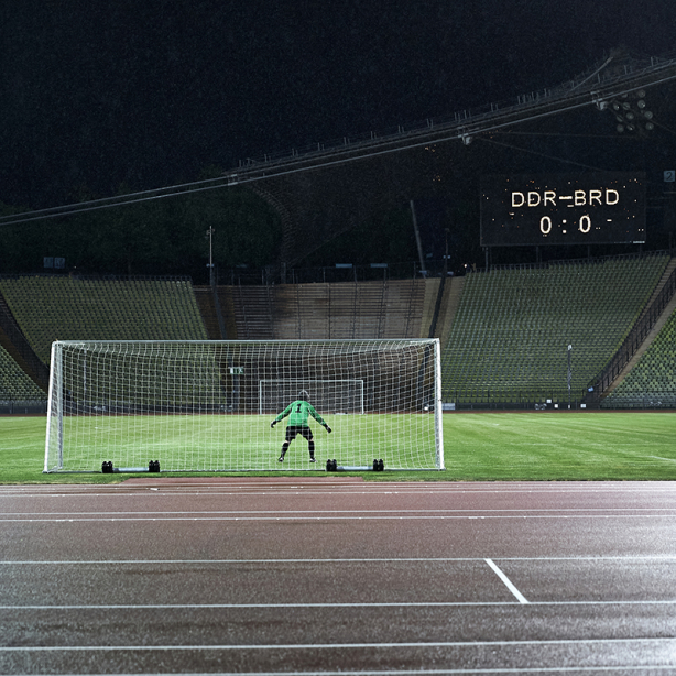 Das Motiv zeigt den Künstler Massimo Furlan im Fußballtrikot in einem Tor im Olympiastadion stehen. Auf einer Anzeigetafel erscheint der Spielstand: "DDR gegen BRD – 0 zu 0".