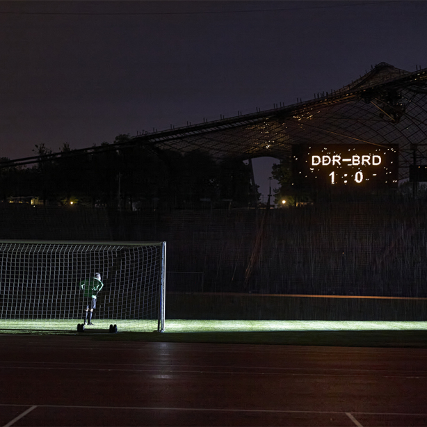 The motif shows the artist Massimo Furlan in a soccer jersey standing in a goal in the Olympic Stadium. On a scoreboard appears the score: "GDR vs FRG - 1 to 0".