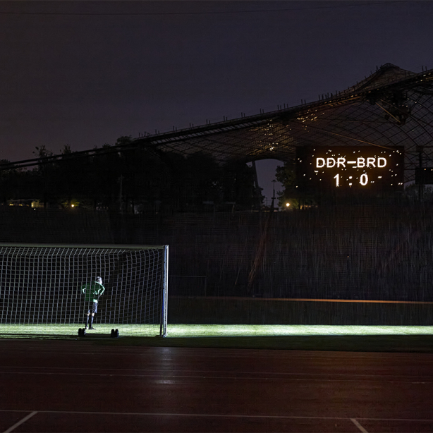 Das Motiv zeigt den Künstler Massimo Furlan im Fußballtrikot in einem Tor im Olympiastadion stehen. Auf einer Anzeigetafel erscheint der Spielstand: "DDR gegen BRD – 1 zu 0".