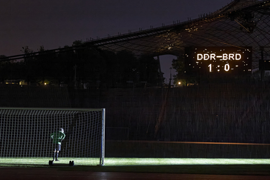 The motif shows the artist Massimo Furlan in a soccer jersey standing in a goal in the Olympic Stadium. On a scoreboard appears the score: "GDR vs FRG - 1 to 0".