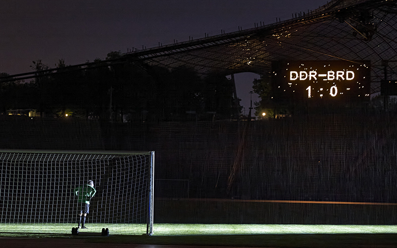 Das Motiv zeigt den Künstler Massimo Furlan im Fußballtrikot in einem Tor im Olympiastadion stehen. Auf einer Anzeigetafel erscheint der Spielstand: "DDR gegen BRD – 1 zu 0".