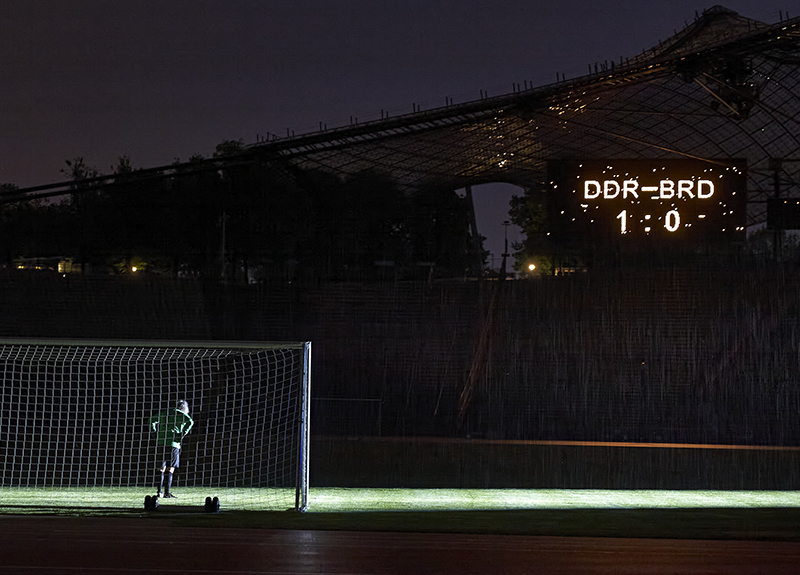 The photograph shows the interior of the Olympic Stadium at night. It shows the artist Massimo Furlan in a green and black football shirt, standing in a goal. The green football turf stretches out in front of him, but he is the only player on the pitch. Only the spotlight of a floodlight on the pitch illuminates the otherwise dark stadium. The score of this fictitious football match appears on a scoreboard high above the spectator stands: "GDR against FRG - 1 to 0".