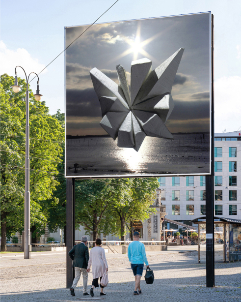 Slight diagonal view of the billboard side facing away from the city center. The motif shows an abstract stone formation floating above the sea in front of a sunset.