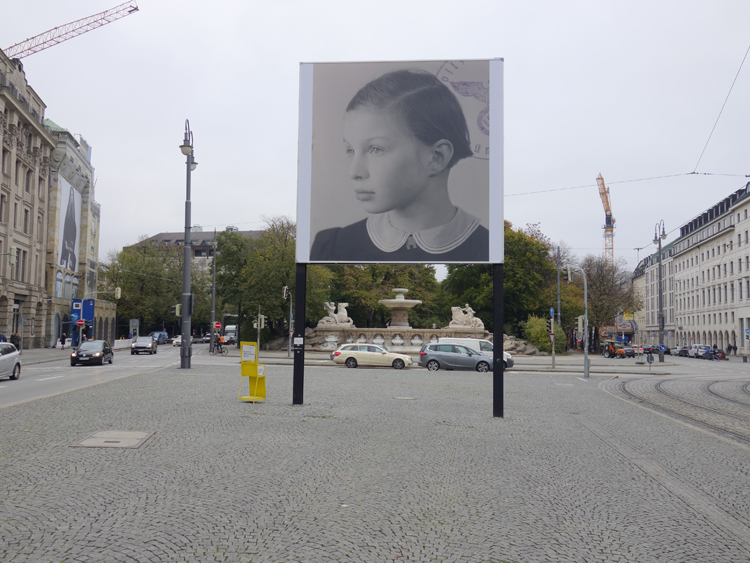 Frontal view of the billboard at Lenbachplatz. The motif shows a black and white photograph of a girl in half-profile. In the right corner appear parts of a round stamp impression, showing the imperial eagle with swastika in an oak wreath in the centre and the lettering "Polizeipräsidium München" (Munich Police Headquarters).