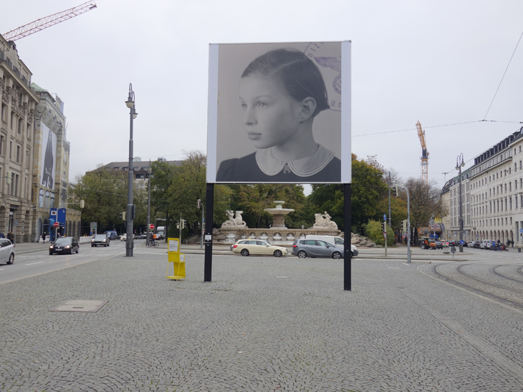 Frontalansicht des Billboards am Lenbachplatz. Das Motiv zeigt die Schwarz-Weiß-Fotografie eines Mädchens im Halbprofil. In der rechten Ecke sind Teile eines runden Stempelabdruckes zu sehen, im Zentrum der Reichsadler mit Hakenkreuz im Eichenkranz sowie der Schriftzug "Polizeipräsidium München".