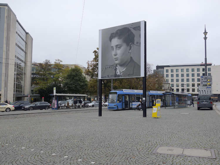 Diagonal view of the billboard at Lenbachplatz. The motif shows a black and white photograph of a boy in half-profile. In the right upper corner and the left lower corner appear parts of a round stamp impression, showing the imperial eagle with swastika in an oak wreath in the centre and the lettering "Polizeipräsidium München" (Munich Police Headquarters).