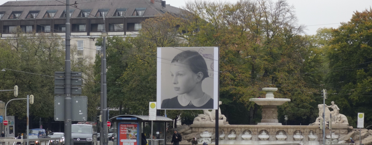 Slightly diagonal view of the billboard at Lenbachplatz. The motif shows a black and white photograph of a girl in half-profile. In the right corner appear parts of a round stamp impression, showing the imperial eagle with swastika in an oak wreath in the centre and the lettering "Polizeipräsidium München" (Munich Police Headquarters).